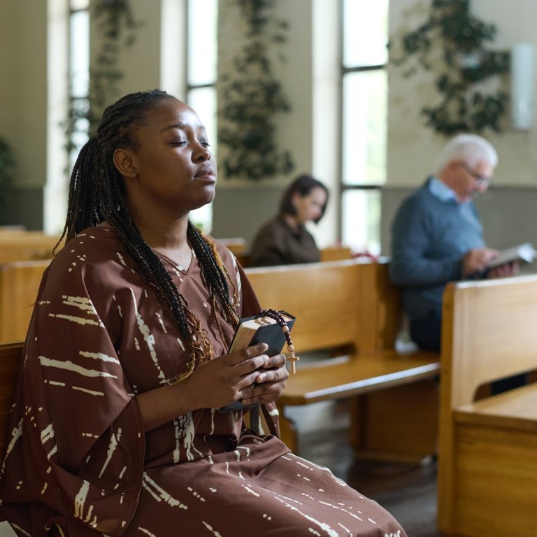 Young Christian black woman in casual dress kkeeping her eyes closed while sitting on bench in church and saying prayers to Lord